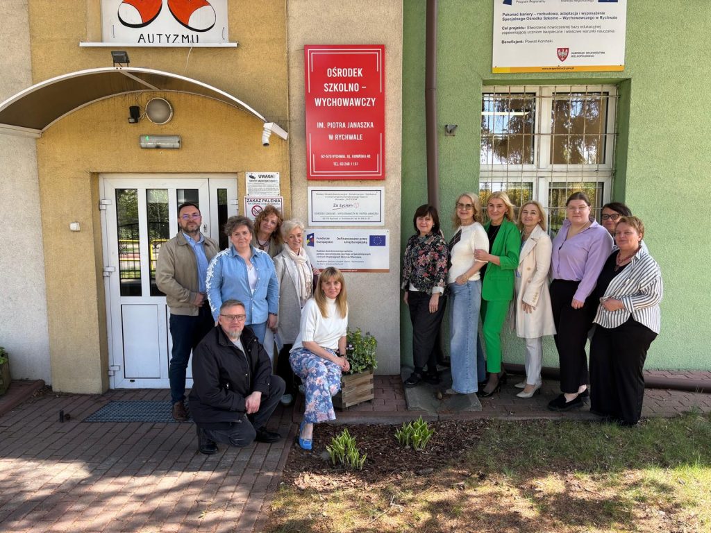 Grupa osób pozuje do zdjęcia przed wejściem do budynku, obok drzwi widać czerwoną tablicę z napisem „Ośrodek Szkolno-Wychowawczy im. Piotra Janaszka w Rychwale” oraz tablice informacyjne, a w tle okno i elewację budynku.