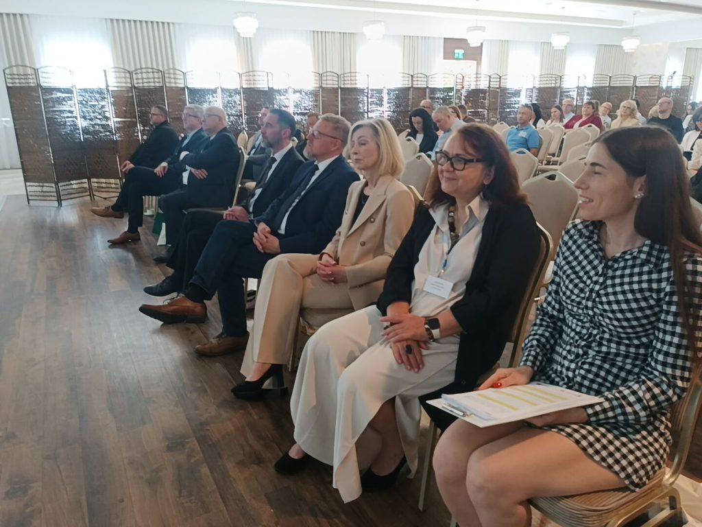 Widownia podczas konferencji. Zgromadzeni uczestnicy siedzą w rzędach na krzesłach w jasnej, przestronnej sali i z uwagą obserwują wydarzenie.