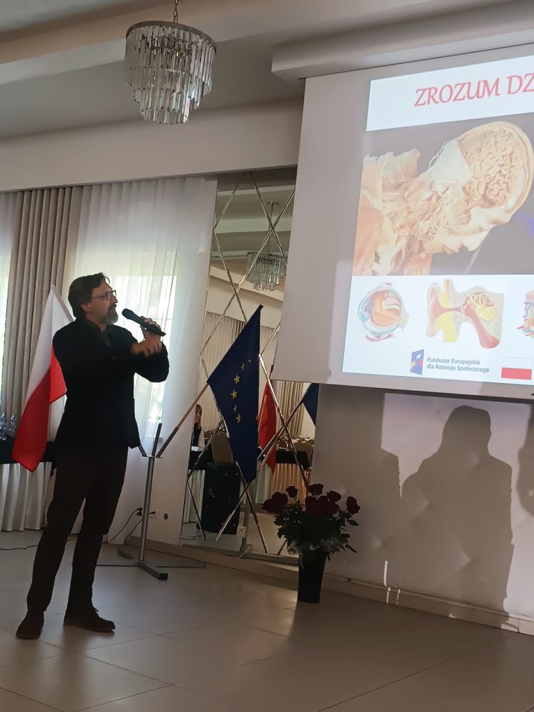 Prelegent z brodą trzyma mikrofon i opowiada o ludzkim organizmie. Ekran w tle wyświetla slajd edukacyjny z anatomicznymi grafikami przedstawiającymi przekroje mózgu, oka i ucha.