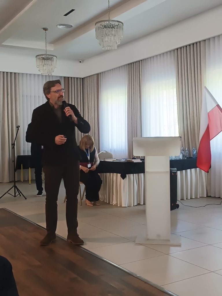 Mężczyzna w okularach i ciemnej marynarce kontynuuje swoje wystąpienie, trzymając mikrofon w dłoni. W głębi sali, tuż obok polskiej flagi, siedzi kobieta przysłuchująca się prelekcji.