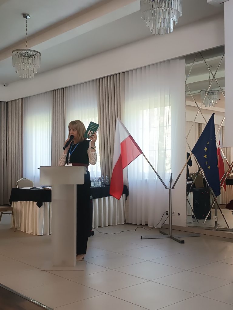 Jasnowłosa kobieta stoi przy białej mównicy i przemawia do publiczności przez mikrofon. Prelegentka prezentuje słuchaczom trzymaną w dłoni zieloną książkę lub broszurę. Tło sceny zdobią flagi Polski oraz Unii Europejskiej.