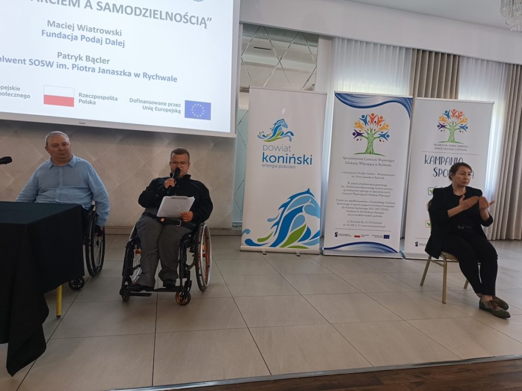 Młody mężczyzna na wózku mówi do mikrofonu podczas konferencji. Obok niego przy stoliku siedzi drugi prelegent, a po prawej stronie kobieta tłumaczy wystąpienie na język migowy. Ekran w tle prezentuje tytuł wystąpienia „Między wsparciem a samodzielnością”.