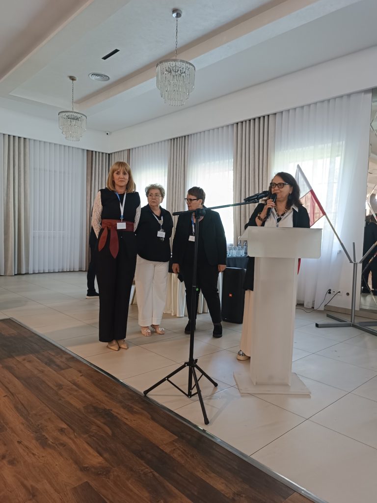 Cztery kobiety stoją na froncie jasnej sali konferencyjnej. Jedna z nich przemawia przez mikrofon przy białej mównicy, podczas gdy pozostałe trzy stoją obok i uważnie przysłuchują się.