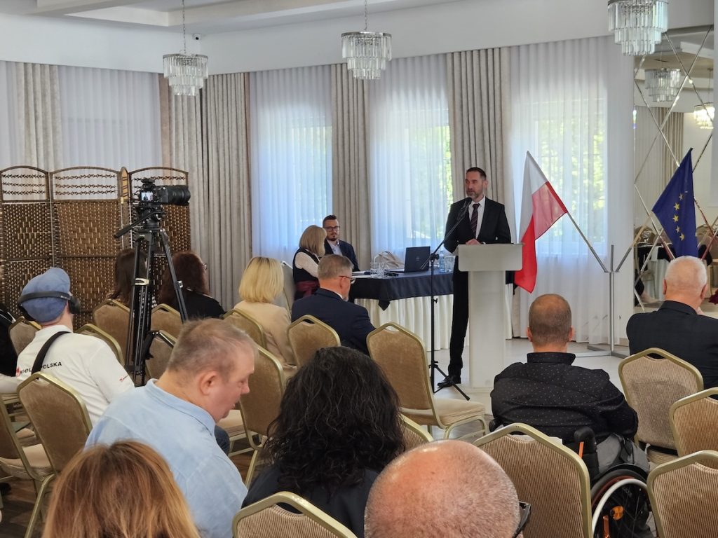 Szeroki kadr sali konferencyjnej, na którym prelegent przemawia zza mównicy stojącej obok flagi Polski i Unii Europejskiej. Uczestnicy siedzą w rzędach, a wydarzenie jest rejestrowane przez kamerę na statywie.