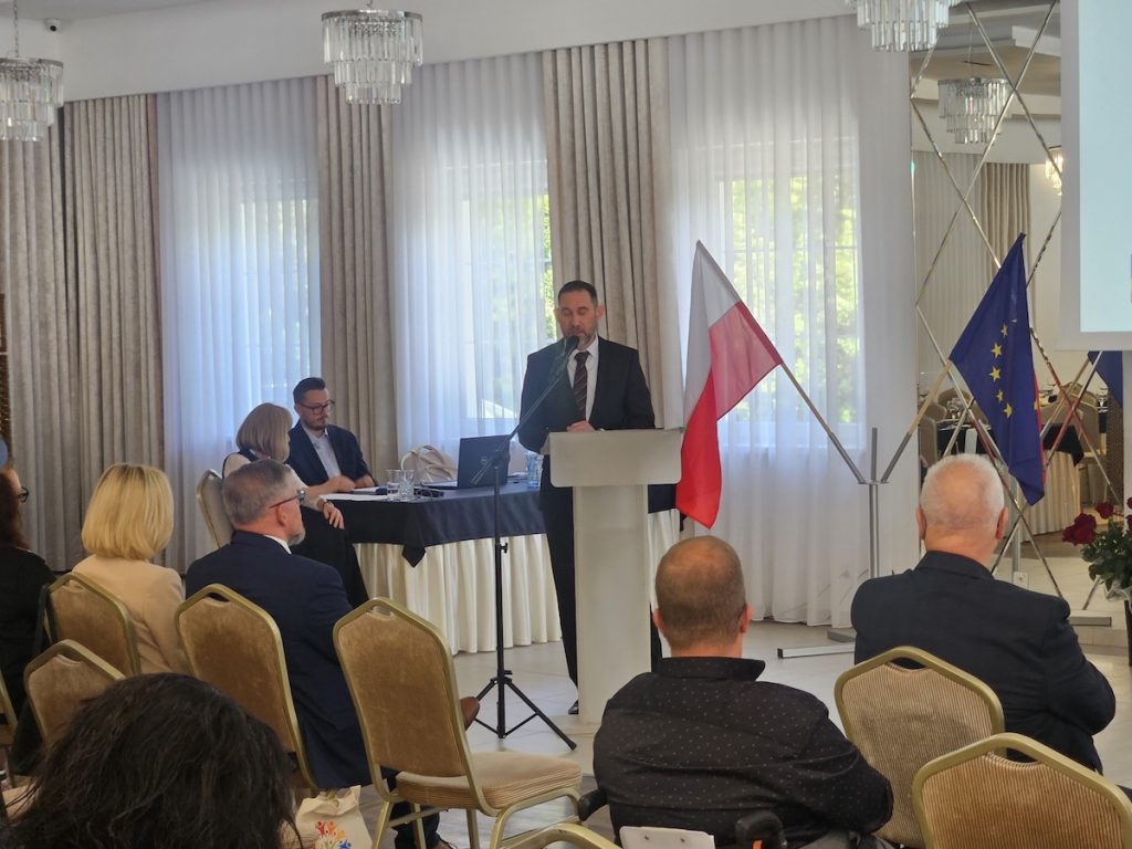 Ujęcie z konferencji, na którym mężczyzna występuje przy mównicy z mikrofonem. W głębi widać inne osoby siedzące za stołem prezydialnym oraz uważnie słuchającą publiczność na pierwszym planie.