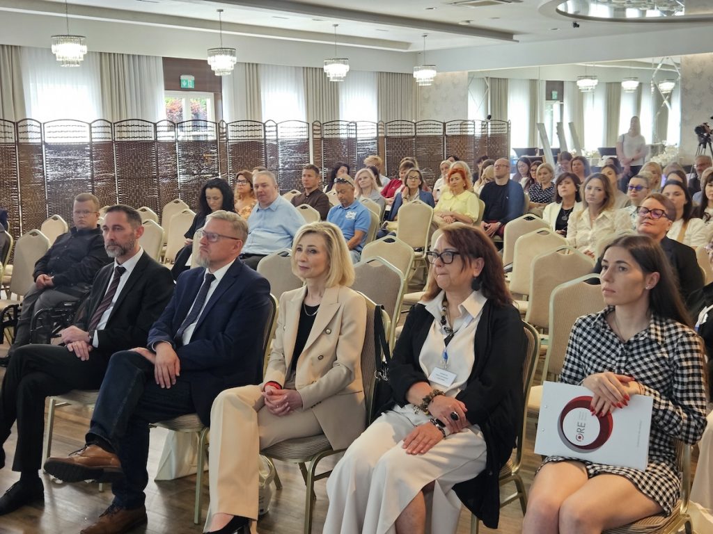 Fotografia przedstawiająca uczestników konferencji siedzących w rzędach i skierowanych w stronę prelegentów. W pierwszym rzędzie widoczna jest między innymi kobieta trzymająca materiały z wyraźnym logo Ośrodka Rozwoju Edukacji (ORE).