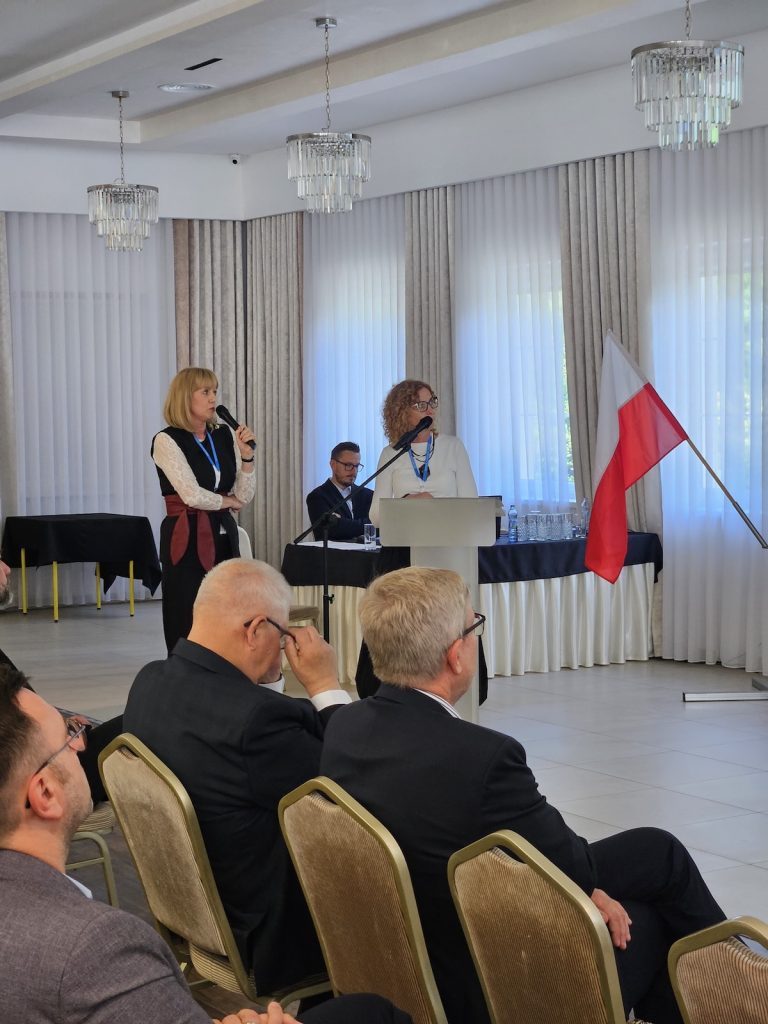 Ujęcie z tego samego wystąpienia, pokazujące dwie kobiety prowadzące prezentację dla zgromadzonych gości. Widoczna na pierwszym planie część publiczności uważnie przysłuchuje się prezentacji.