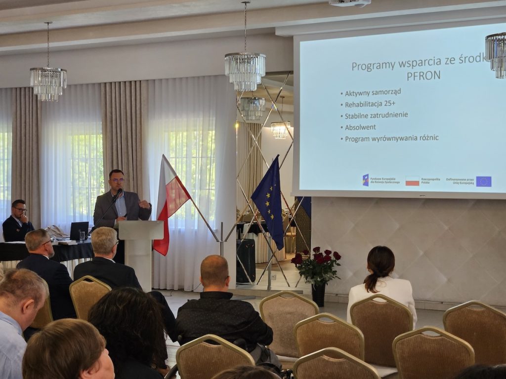 Mężczyzna w garniturze przemawia zza mównicy. Ekran obok niego wymienia programy wsparcia z funduszy PFRON, na przykład „Aktywny samorząd” oraz „Rehabilitacja 25+”. Goście przysłuchują się jego słowom.