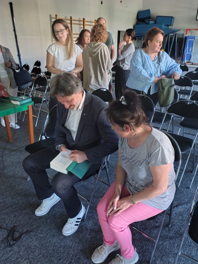 Robert Bielecki siedzi i daje autograf w książce. Obok na krześle czeka kobieta w różowych spodniach. Z tyłu w sali gimnastycznej stoją i rozmawiają inne osoby.