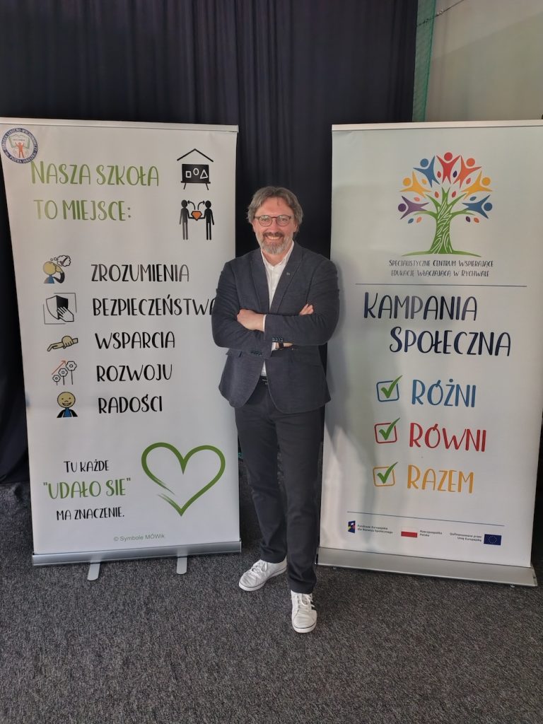 Robert Bielecki stoi z rękami na piersi między dwoma informacyjnymi roll-upami. Baner po lewej ma napis: Nasza szkoła to miejsce. Baner po prawej promuje kampanię społeczną Różni – Równi – Razem.