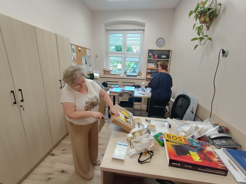 Kobieta na pierwszym planie otwiera nożykiem małe pudełko na biurku pełnym materiałów. W głębi druga kobieta pracuje przy stole pod oknem.