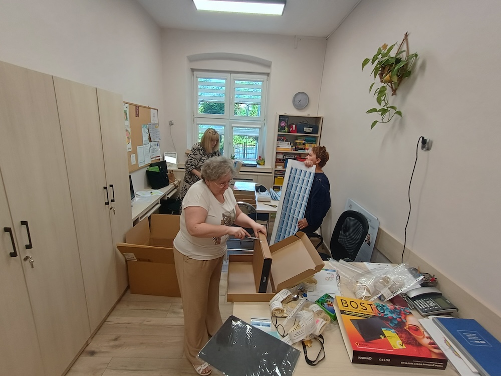 Kobieta przy biurku rozcina nożykiem duży, płaski karton. Z tyłu druga kobieta trzyma duży arkusz naklejek, a trzecia stoi przy oknie.
