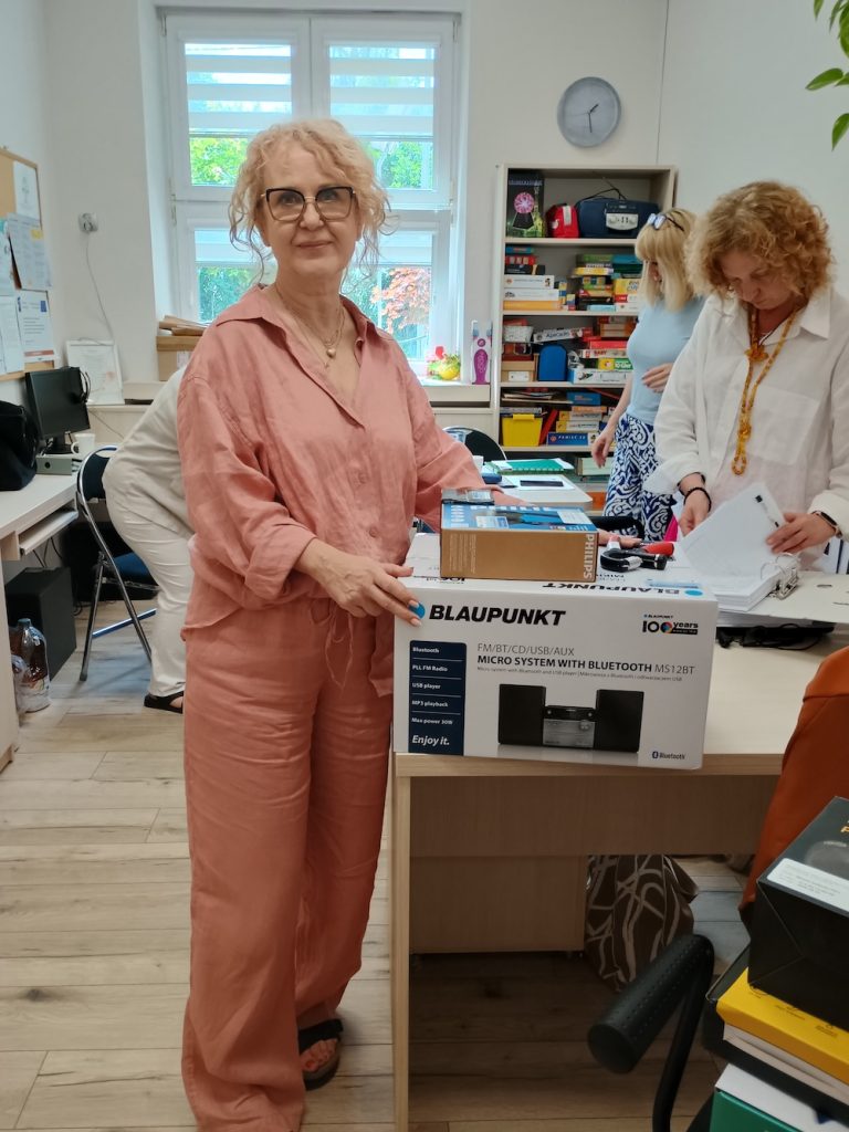 Kobieta w różowym, lnianym komplecie opiera dłonie na kartonie ze sprzętem audio marki Blaupunkt. Za nią w głębi biura inna kobieta w białej koszuli porządkuje dokumenty.