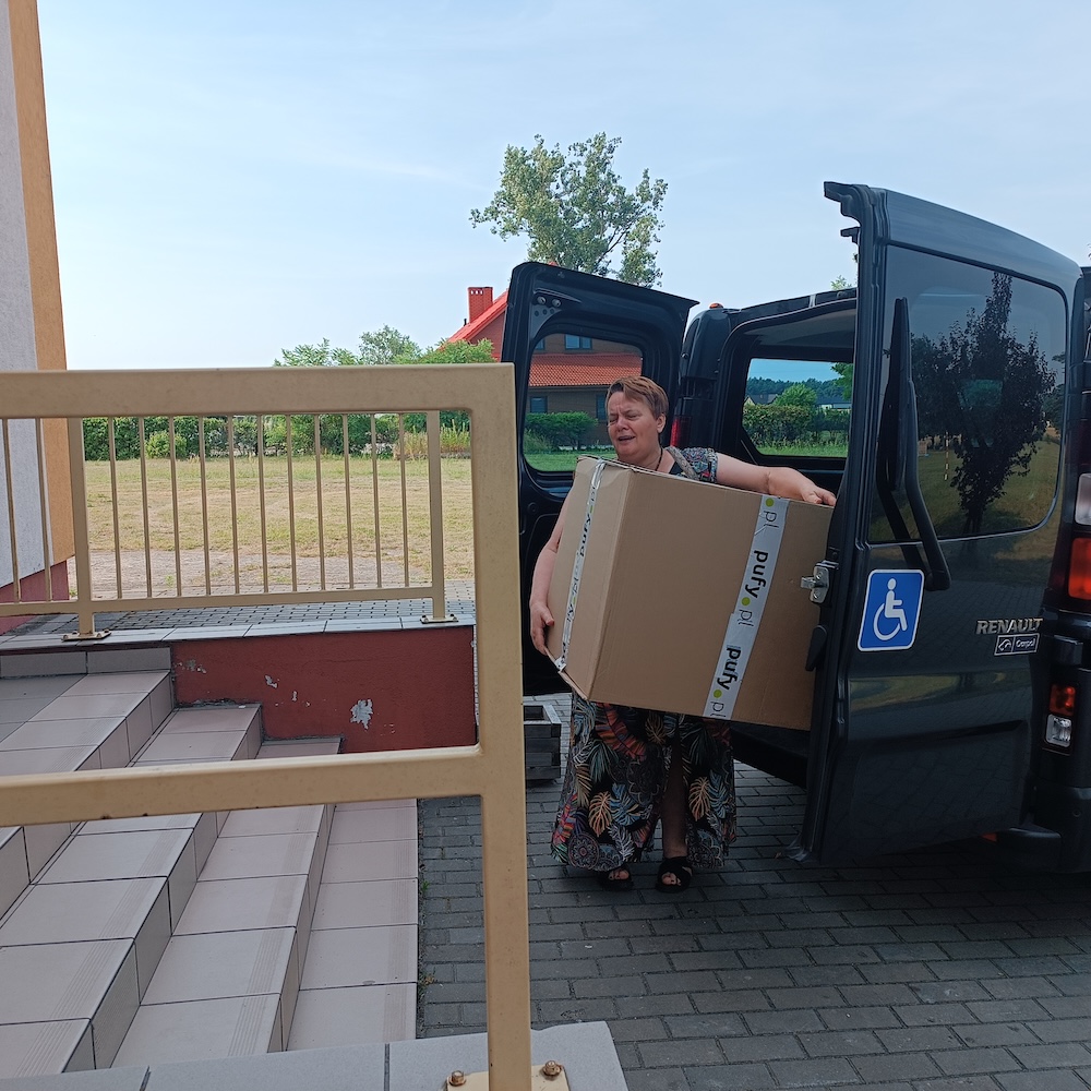 Kobieta w kolorowej sukience niesie duży karton. Wyjmuje paczkę z bagażnika ciemnego busa. Pojazd stoi tuż przy schodach do budynku.